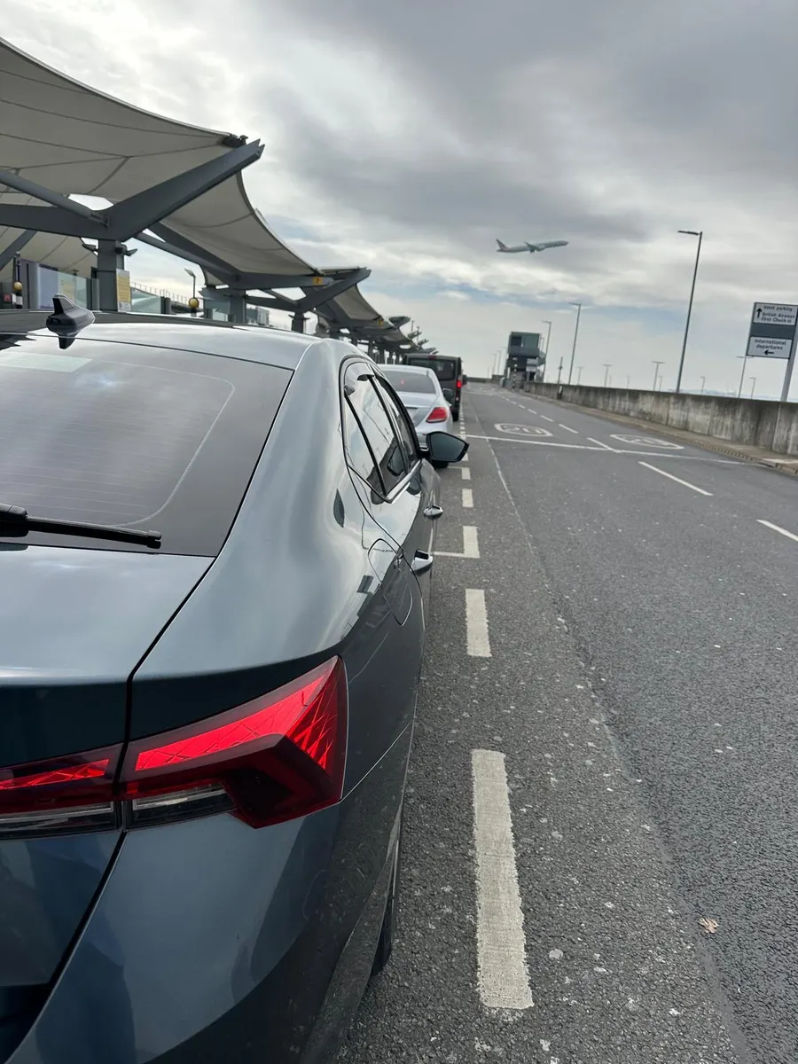 Grey car at an airport drop-off with a plane overhead